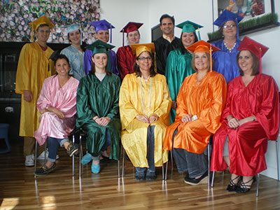 Group of graduates in colorful robes and caps seated and standing indoors at graduation ceremony.