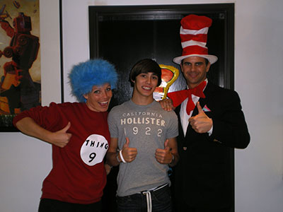 Three people in costume pose together indoors: one wearing blue wig, one in Hollister shirt, one in top hat and tuxedo.