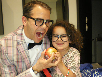 Man and child wearing glasses hold a red apple together indoors, smiling playfully.