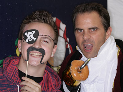 Two men at party wearing pirate and casual costumes, holding food items indoors.