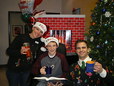Three people wearing Santa hats pose together holding a book in front of a decorated fireplace during holiday celebration.