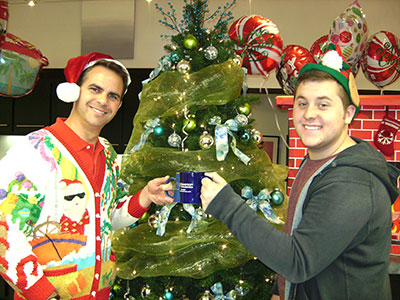 Two men wearing Santa hats decorate a Christmas tree in an office setting.
