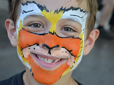 Child with face painted as tiger, smiling at camera in indoor setting.
