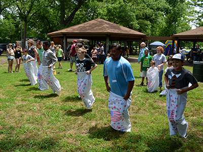 Children in colorful outfits participate in outdoor community event under brown umbrellas on grass.