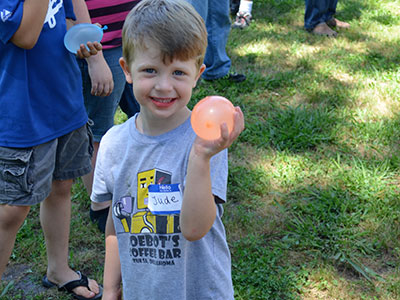 Young child holding a peach at outdoor event, wearing gray shirt with blue sleeve nearby.