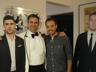 Four men posing together indoors, two wearing formal attire and two in casual clothing at what appears to be a social event.