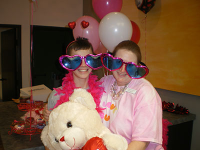 Two children wearing colorful heart-shaped glasses hold a large white teddy bear at a Valentine's Day party.