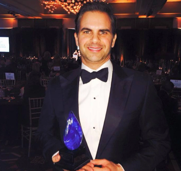 Man in formal tuxedo holding blue award at elegant indoor event venue.