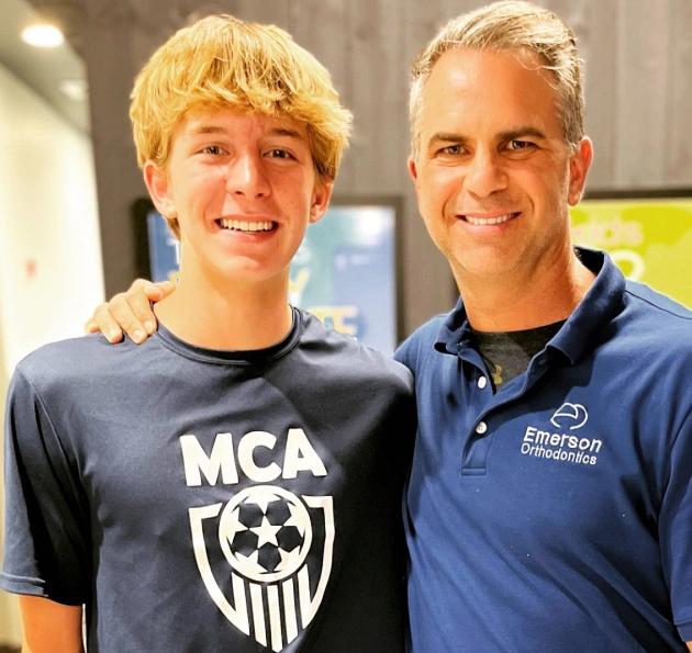 Two people smiling together at Emerson Orthodontics clinic, one wearing MCA shirt, one in blue polo.