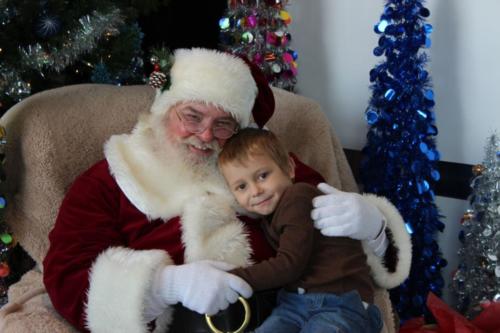 Santa Claus holding young child on lap in festive holiday setting with decorated trees.