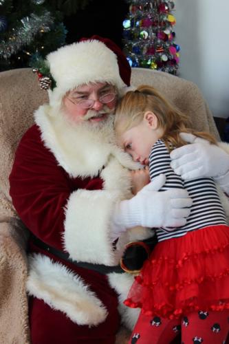 Santa Claus holds smiling toddler on lap in festive holiday setting with Christmas tree.