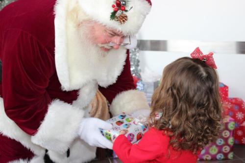 Santa Claus giving a gift to a young girl in red dress during holiday celebration.