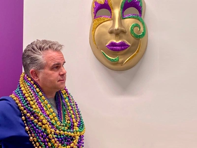 Man wearing Mardi Gras beads stands beside colorful theatrical mask on wall.
