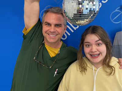 Two people smiling at camera in front of blue backdrop with disco ball at orthodontics event.