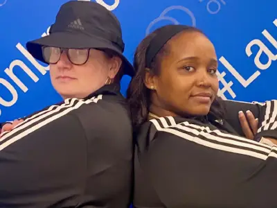 Two people in black Adidas jackets pose back-to-back against blue branded backdrop at event.