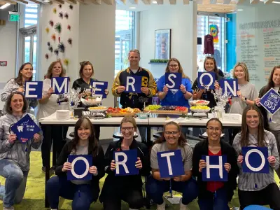 Emerson Orthodontics team members wearing blue shirts spell out "EMERSON ORTHO" in an office setting.