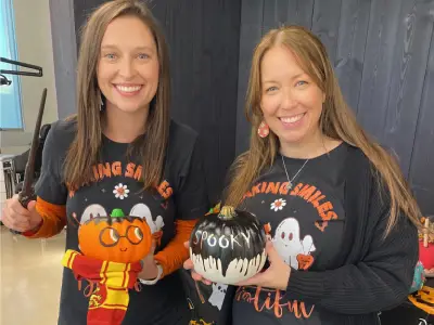 Two women wearing Halloween sweatshirts smile at camera in office setting.