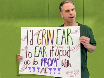 Man holding handmade sign about wearing ear gear for prom, green background, orthodontics promotion.
