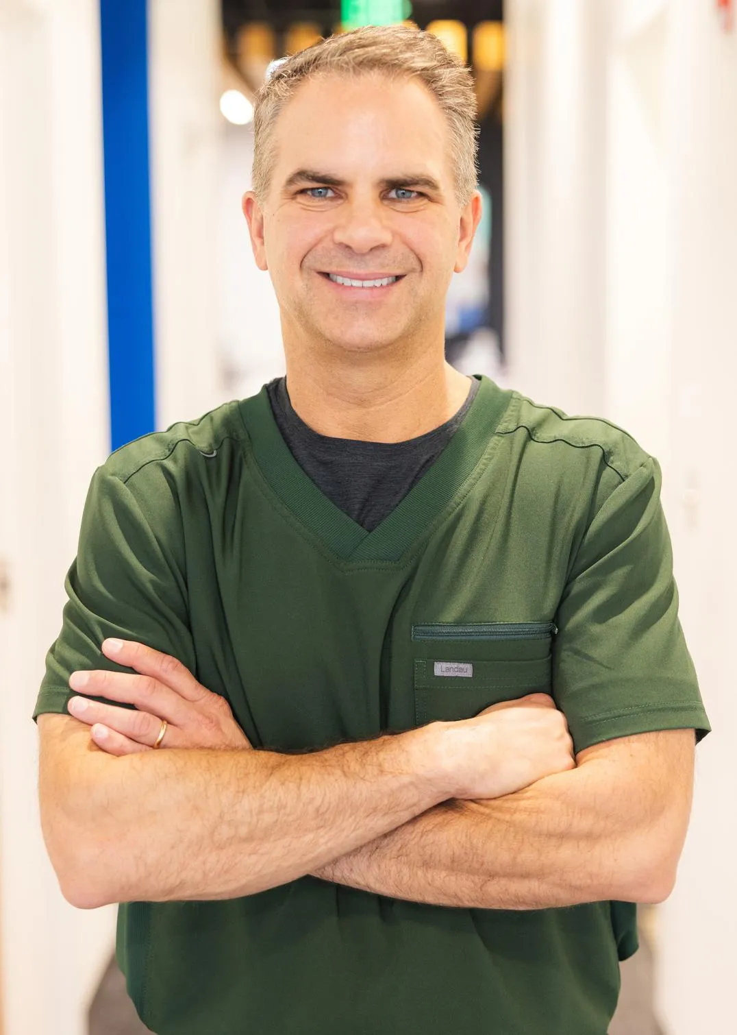 Male orthodontist in green scrubs smiling with arms crossed in clinical setting.