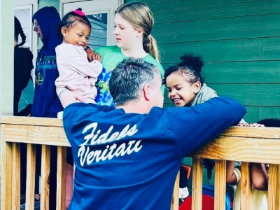 Man in blue jacket holding child while interacting with woman and child on wooden porch.