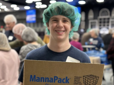 Young man wearing hairnet holds MannaPack box at community event or food bank.