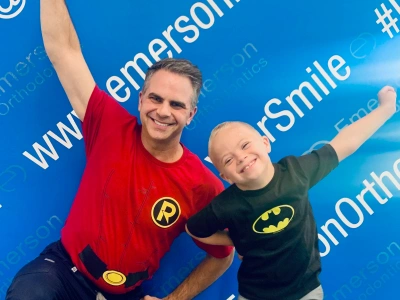 Adult and child wearing superhero shirts pose happily against blue Emerson Orthodontics branded backdrop.