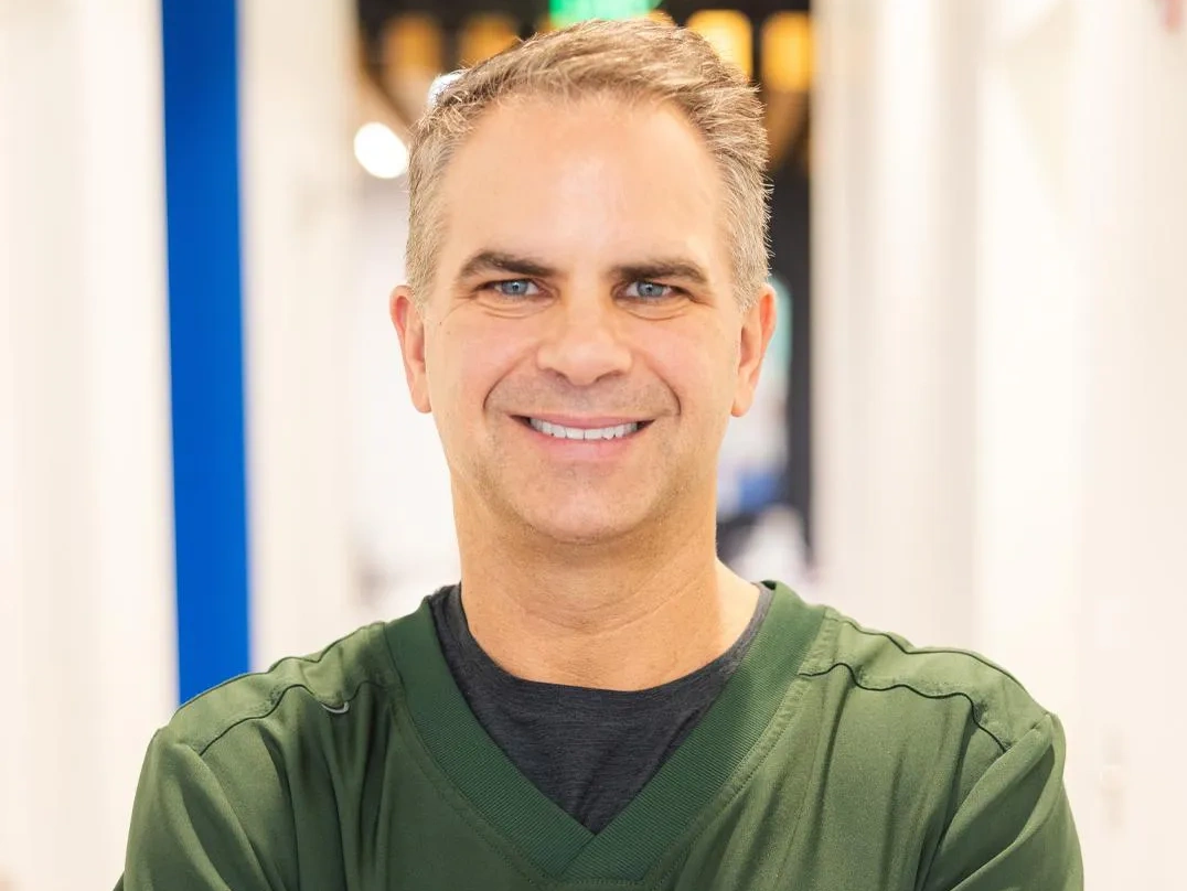 Smiling male orthodontist wearing olive green shirt in modern dental office setting.