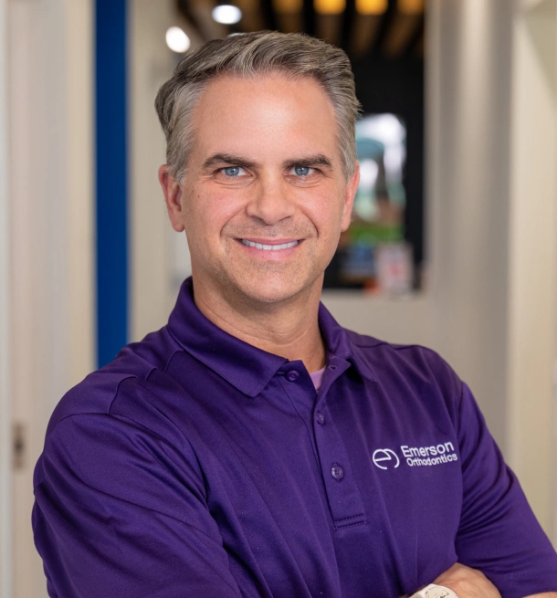 miling male orthodontist with graying hair wearing a purple Emerson Orthodontics polo shirt, standing with arms crossed in an office hallway.