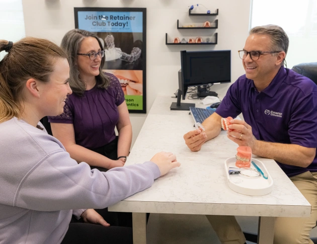 An orthodontist uses a dental model to explain a treatment plan to a female patient and her mother at a consultation desk.